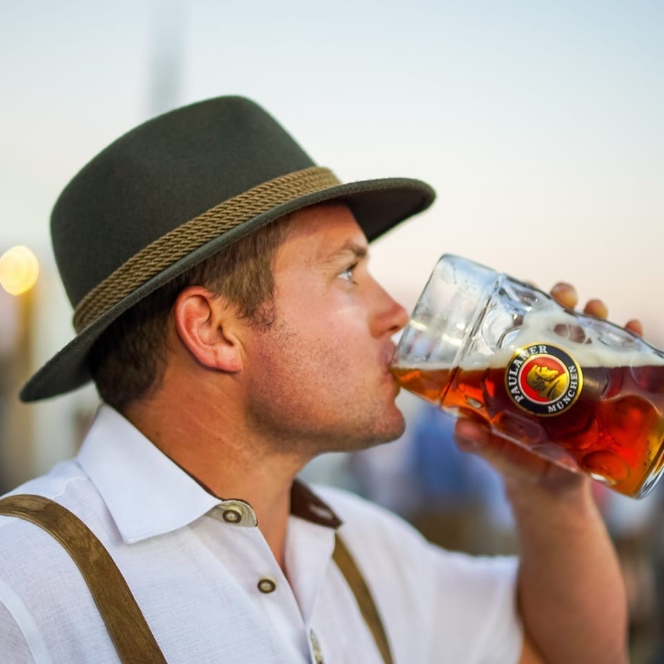 Man in a hat and suspenders drinking from a large glass of beer.