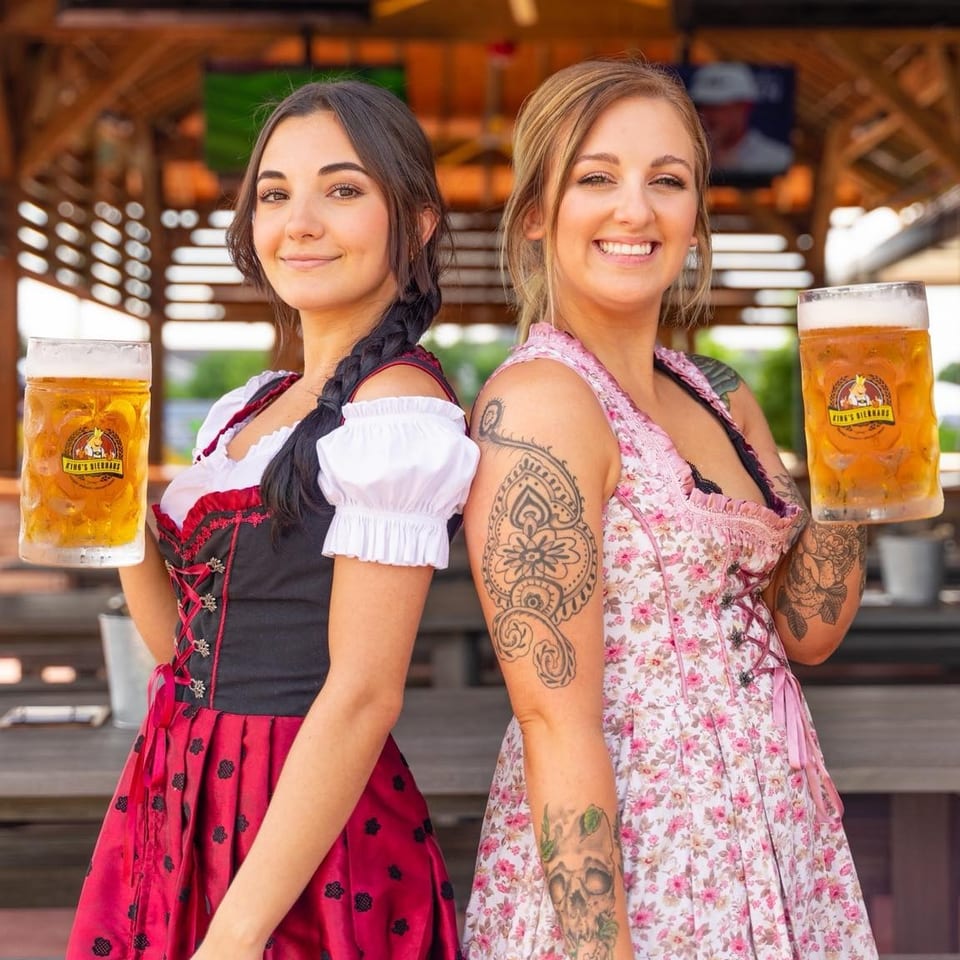Two smiling women in dirndls hold large steins of beer at King's Bierhaus.