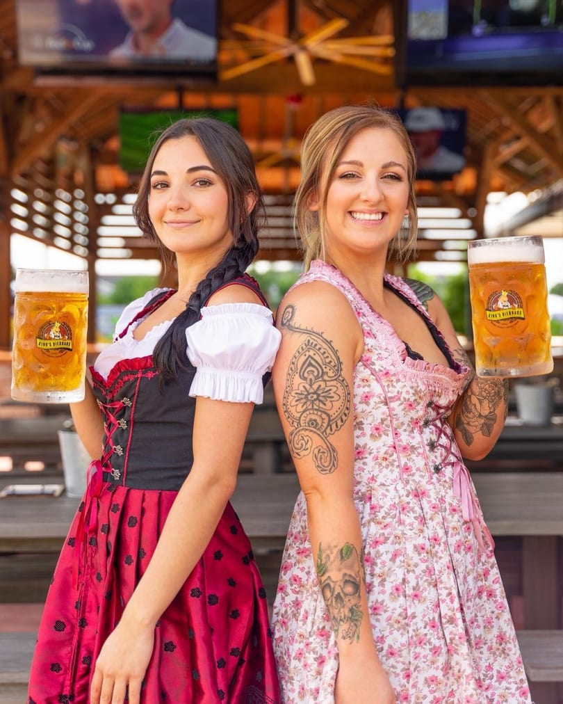 Two smiling women in dirndls hold large steins of beer at King's Bierhaus.