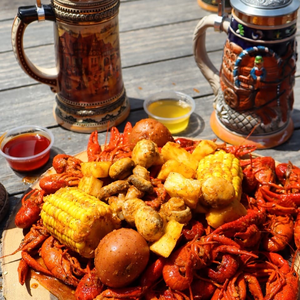 Boiled crawfish, corn, potatoes, and mushrooms on a wooden board with two beer steins.