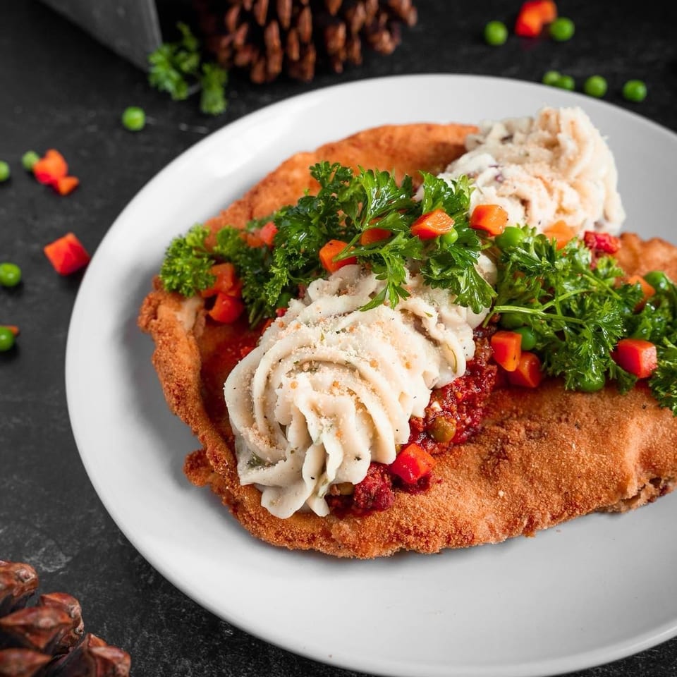 Crispy breaded cutlet topped with mashed potatoes, red sauce, peas, carrots, and parsley on a white plate.