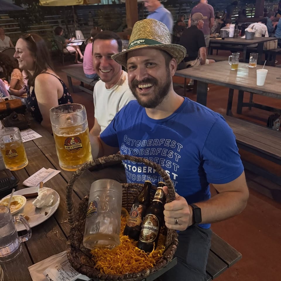 Man in Oktoberfest shirt and straw hat holding a beer basket, smiling at a lively outdoor table.