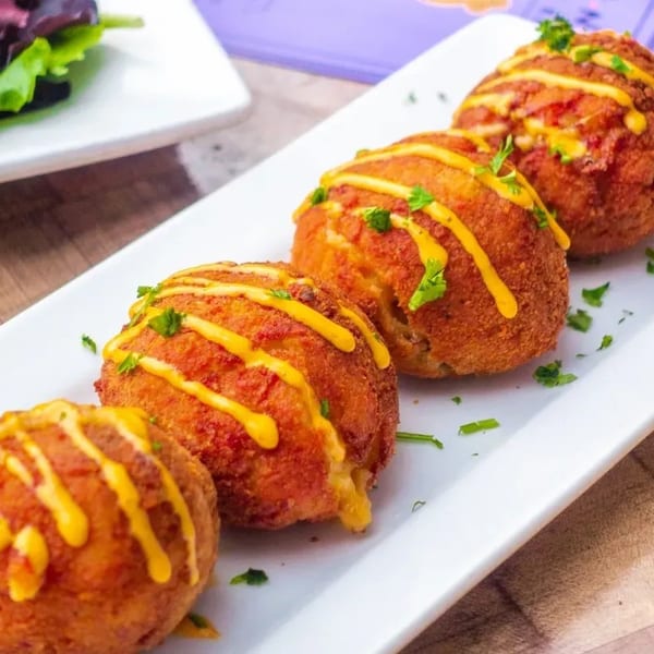 Four golden-brown fried arancini balls drizzled with yellow sauce and garnished with parsley on a white plate.