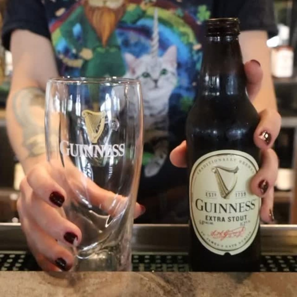 A person holding a Guinness beer bottle and an empty Guinness glass on a bar.