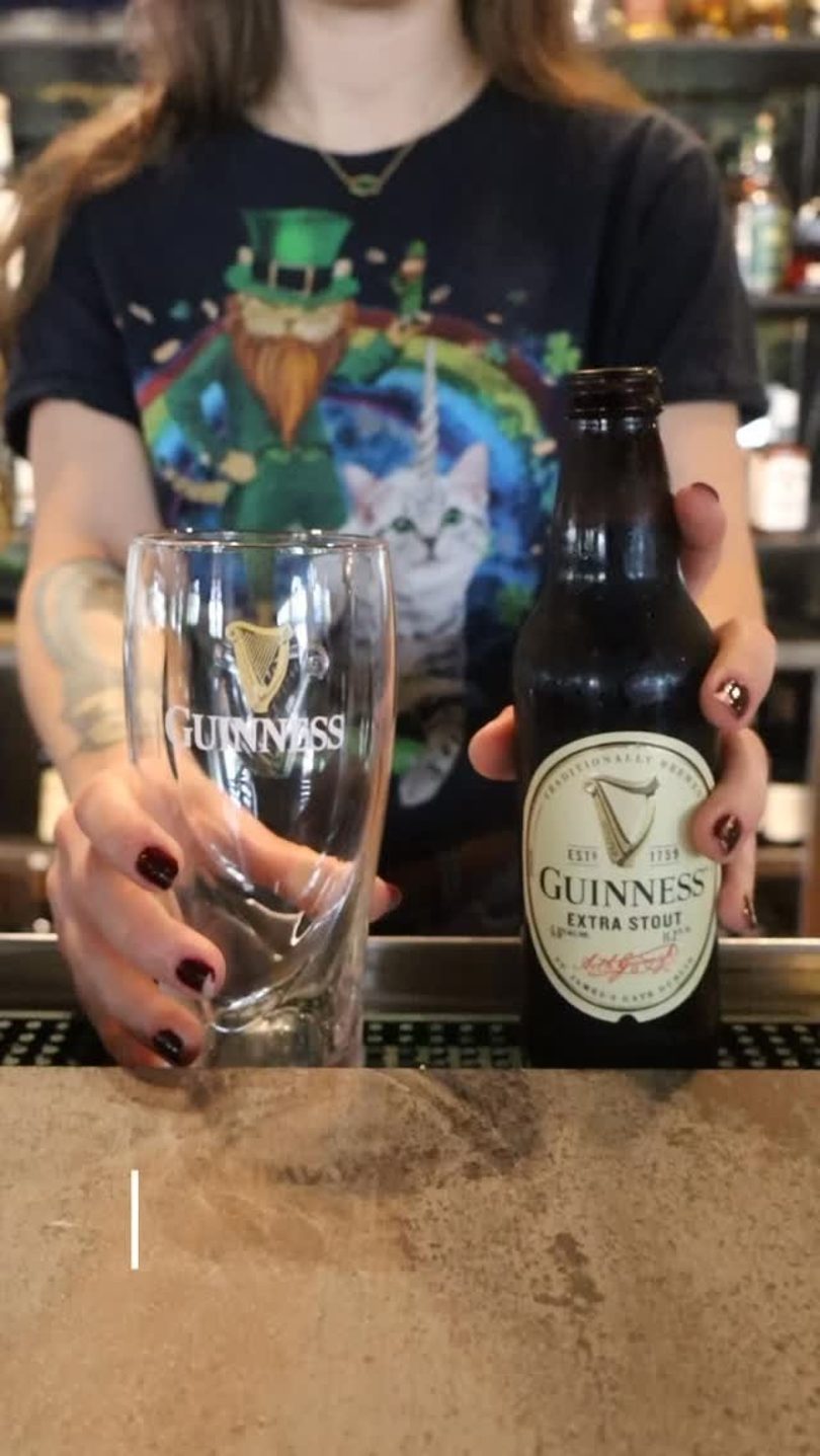 A person holding a Guinness beer bottle and an empty Guinness glass on a bar.