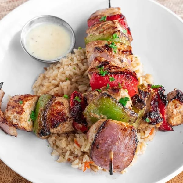 Two chicken and vegetable skewers with rice and a side of sauce on a white plate.
