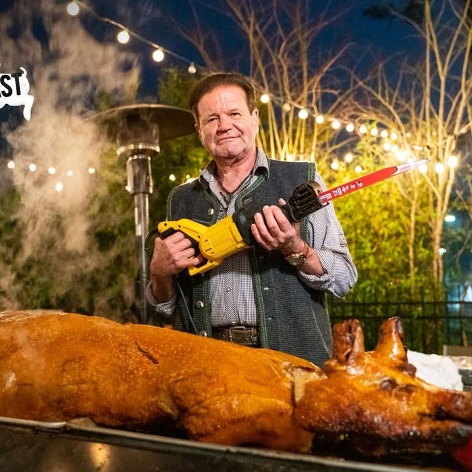 Man in Bavarian attire holding a saw next to a roasted pig with an apple in its mouth.