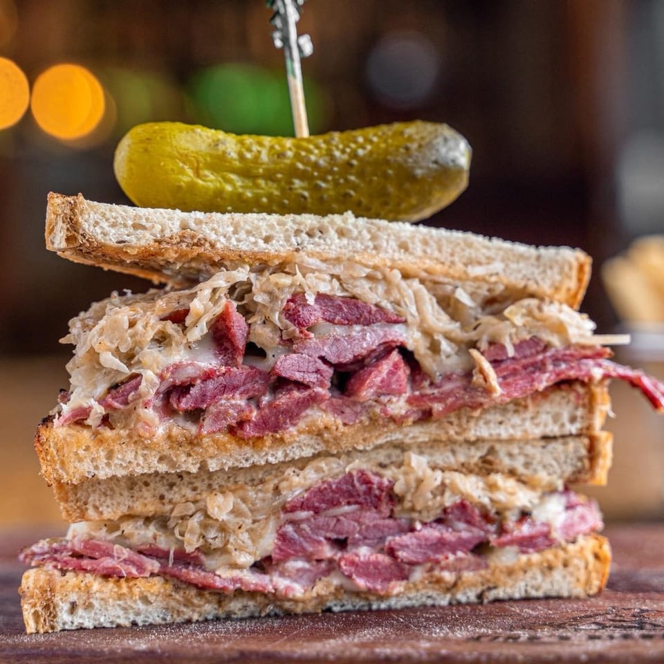 Stacked pastrami and sauerkraut sandwich on toasted bread, with a pickle spear on top.
