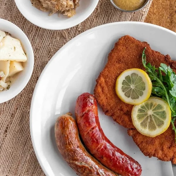 A plate of schnitzel with lemon slices and arugula, two sausages, and sides of potato salad and sauerkraut.