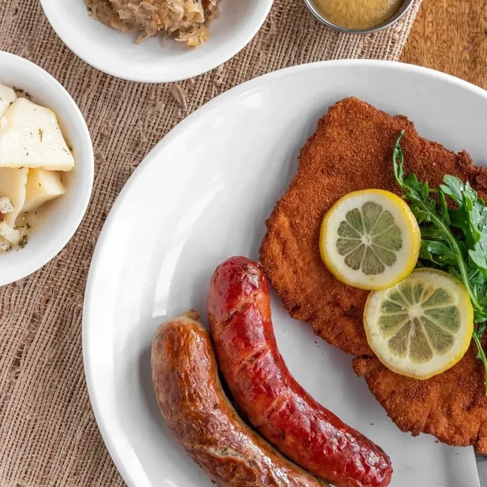 A plate of schnitzel with lemon slices and arugula, two sausages, and sides of potato salad and sauerkraut.