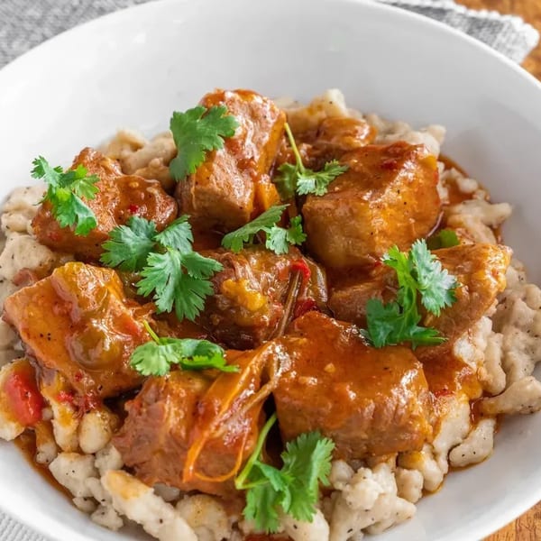 Hearty goulash with tender meat and dumplings, garnished with fresh cilantro in a white bowl.