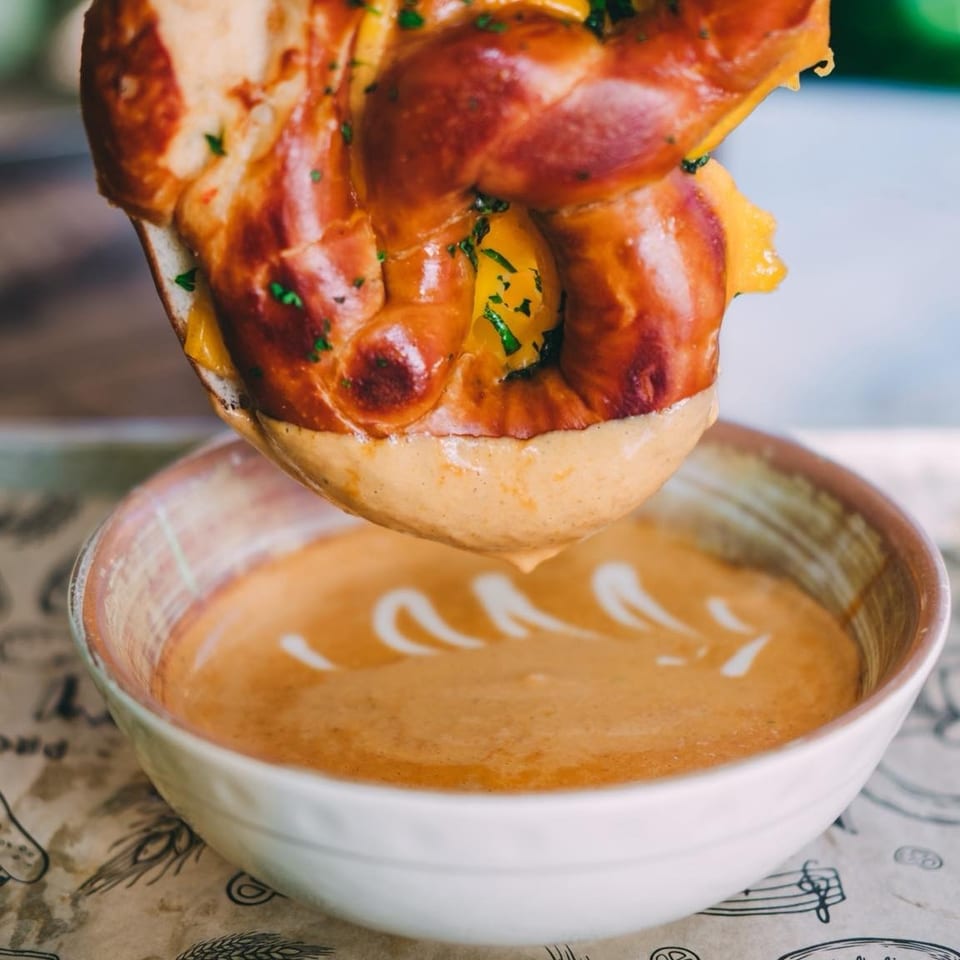 A cheesy pretzel being dipped into a bowl of creamy orange soup with white swirls.