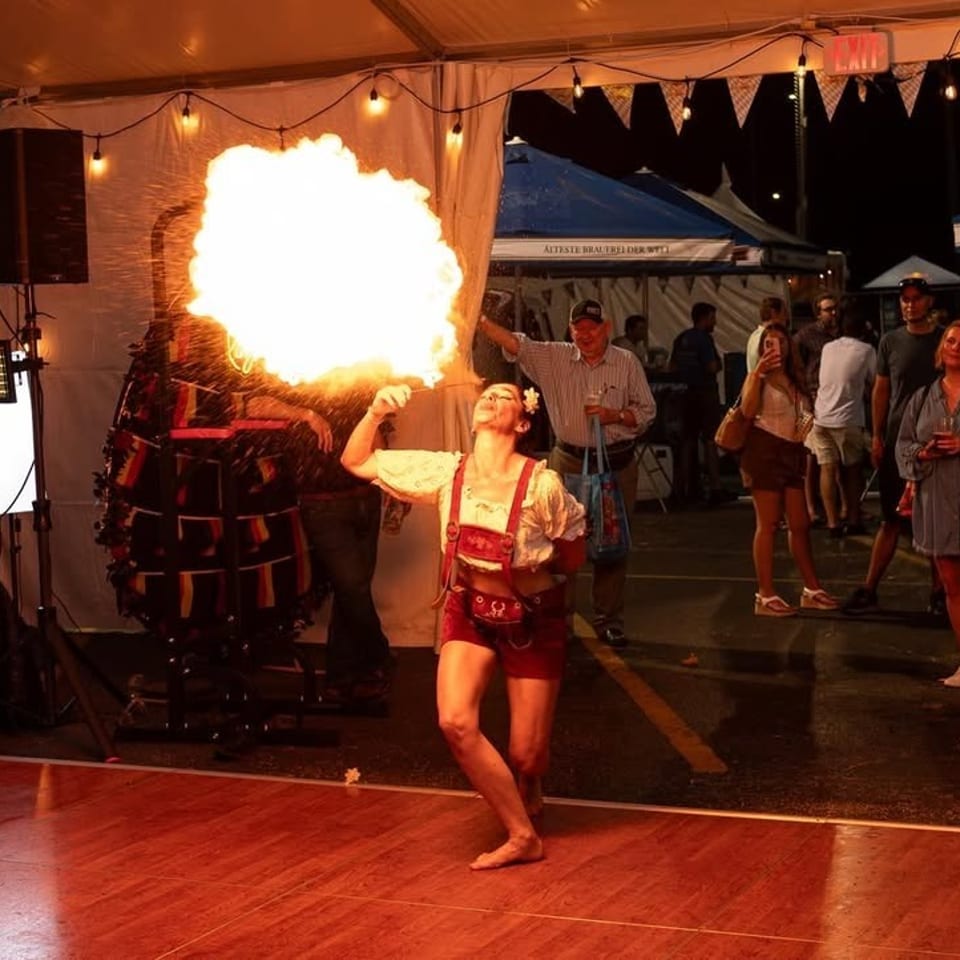 A woman in traditional German attire breathes fire on a dance floor, entertaining a crowd at night.