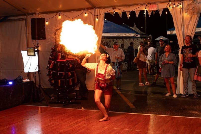 A woman in traditional German attire breathes fire on a dance floor, entertaining a crowd at night.